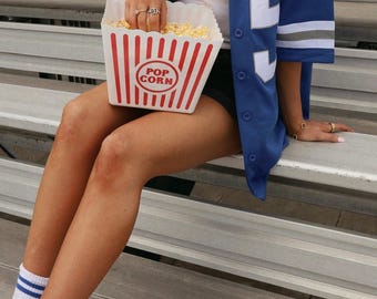 May include: A blue button-down jersey with white and grey stripes on the sleeves and a white number on the front, worn over a white tank top with black shorts. White crew socks with two blue stripes and grey athletic sneakers are worn with the outfit.