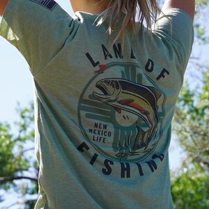 May include: A green t-shirt with a graphic of a fish jumping out of the water. The graphic is surrounded by a circle with the text "Land of New Mexico Life Fishing".