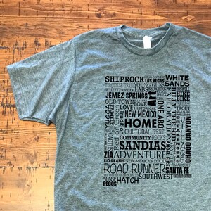 May include: A heather grey t-shirt with a black graphic print of a word cloud featuring places and things related to New Mexico. The words include "Shiprock", "Las Vegas", "White Sands", "Taos", "Jemez Springs", "Old Town", "Love", "New Mexico", "Home", "Cultural", "Community", "Sandals", "Adventure", "Rio Grande", "Road Runners", "Santa Fe", "Hatch", "Southwest", and more.