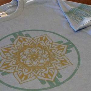 Peut inclure: Un t-shirt bleu clair avec un motif de mandala doré et vert. Le mandala est un motif circulaire avec une fleur au centre et des motifs complexes qui rayonnent vers l'extérieur.