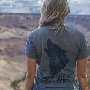 Peut inclure: Une femme portant un t-shirt gris avec un loup noir imprimé et le texte "Stay Wild" sur le dos. Elle se tient devant une vue panoramique d'un canyon.