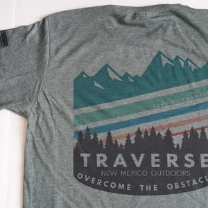 May include: Gray t-shirt with a graphic design of mountains, trees, and stripes in shades of blue, green, and red. The text "Traverse New Mexico Outdoors Overcome The Obstacle" is printed on the shirt.