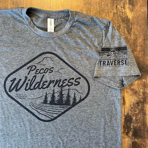 May include: Gray heather t-shirt with a black graphic of a mountain range and the text "Pecos Wilderness" and "Santa Fe National Forest". The right sleeve has a black patch with the text "Traverse" and a mountain range graphic.