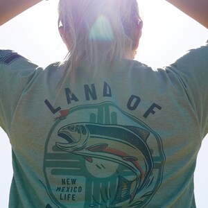 May include: A green t-shirt with a graphic of a fish jumping out of the water. The graphic is surrounded by a circle with the text "Land of Fishing" and "New Mexico Life".