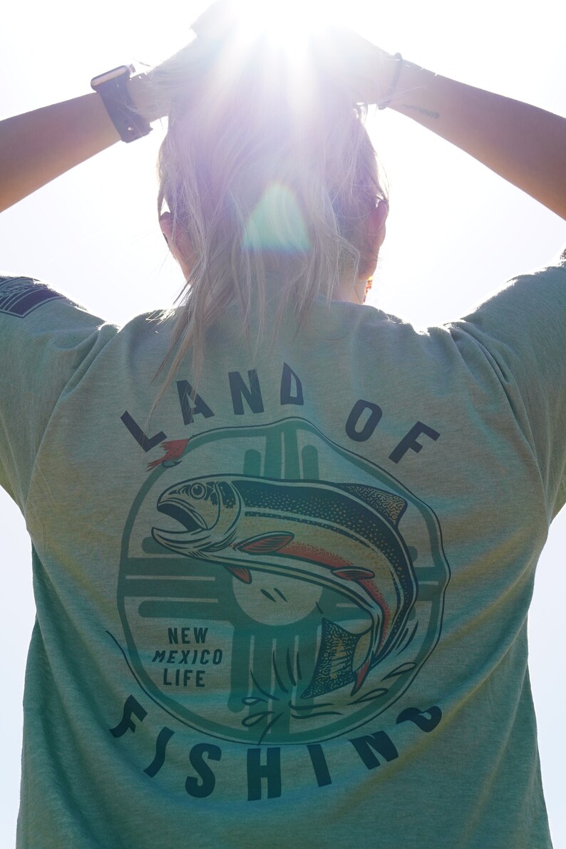 May include: A green t-shirt with a graphic of a fish jumping out of the water. The graphic is surrounded by a circle with the text "Land of Fishing" and "New Mexico Life".