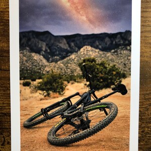 Pode incluir: Uma bicicleta de montanha preta com detalhes amarelos est&aacute; deitada de lado em um caminho de terra. A bicicleta est&aacute; na frente de uma cordilheira com um c&eacute;u estrelado acima.