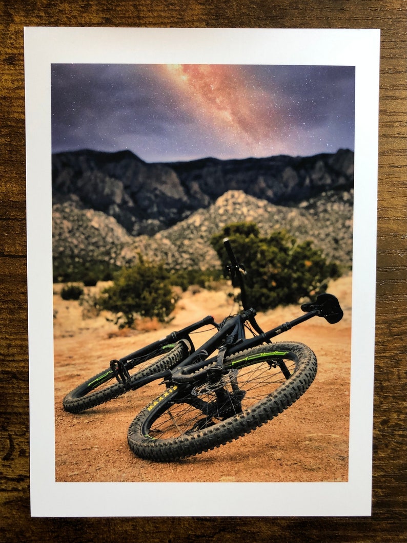Pode incluir: Uma bicicleta de montanha preta com detalhes amarelos est&aacute; deitada de lado em um caminho de terra. A bicicleta est&aacute; na frente de uma cordilheira com um c&eacute;u estrelado acima.