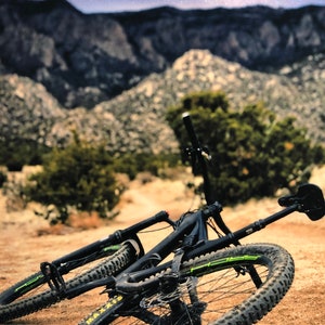Pode incluir: Uma bicicleta de montanha preta com detalhes verdes est&aacute; deitada no ch&atilde;o. A bicicleta est&aacute; na frente de uma cordilheira com um c&eacute;u estrelado ao fundo.
