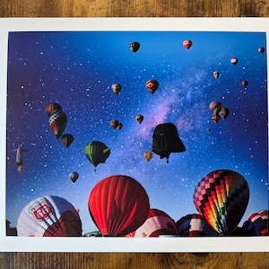May include: A colorful hot air balloon festival with a dark silhouette of Darth Vader in the sky. The balloons are various colors and shapes, including a red balloon with "Route 66" printed on it. The sky is a deep blue with a milky way galaxy in the background.