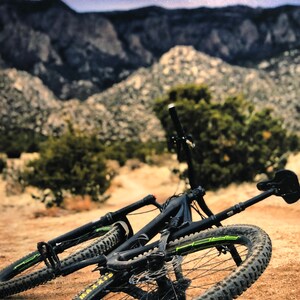 Pode incluir: Uma bicicleta de montanha preta com detalhes verdes est&aacute; deitada de lado em um caminho de terra. A bicicleta tem um garfo dianteiro com suspens&atilde;o e um amortecedor traseiro. A bicicleta est&aacute; na frente de uma cordilheira com um c&eacute;u estrelado no fundo.