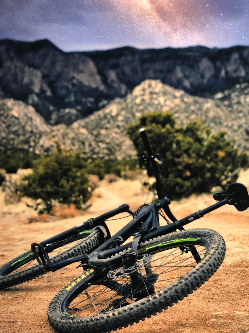 Pode incluir: Uma bicicleta de montanha preta com detalhes verdes est&aacute; deitada de lado em um caminho de terra. A bicicleta tem um garfo dianteiro com suspens&atilde;o e um amortecedor traseiro. A bicicleta est&aacute; na frente de uma cordilheira com um c&eacute;u estrelado no fundo.