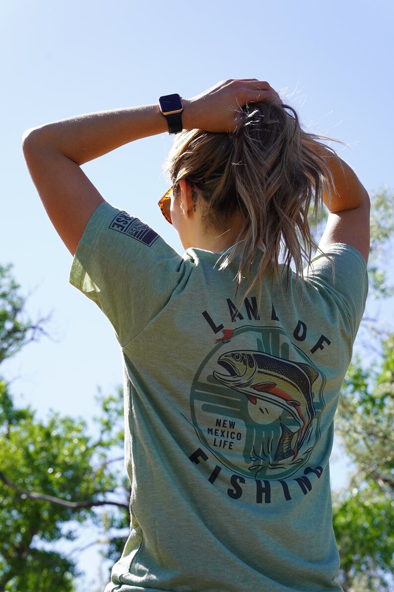 May include: A green t-shirt with a graphic of a fish jumping out of the water. The graphic is surrounded by a circle with the text "Land of New Mexico Life Fishing".