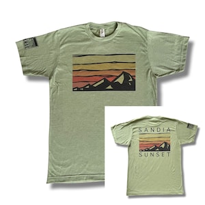 Könnte beinhalten: Ein grünes T-Shirt mit einem Aufdruck einer Bergkette und einem Sonnenuntergang. Auf der Rückseite des Shirts steht "Sandia Sunset".