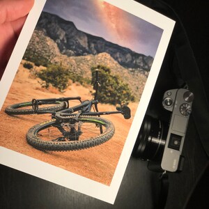 Pode incluir: Uma bicicleta de montanha preta com detalhes verdes est&aacute; deitada no ch&atilde;o em uma paisagem des&eacute;rtica. A bicicleta tem suspens&atilde;o dianteira e pneus com cravos. O fundo mostra uma cordilheira e um c&eacute;u nublado com um toque da Via L&aacute;ctea.