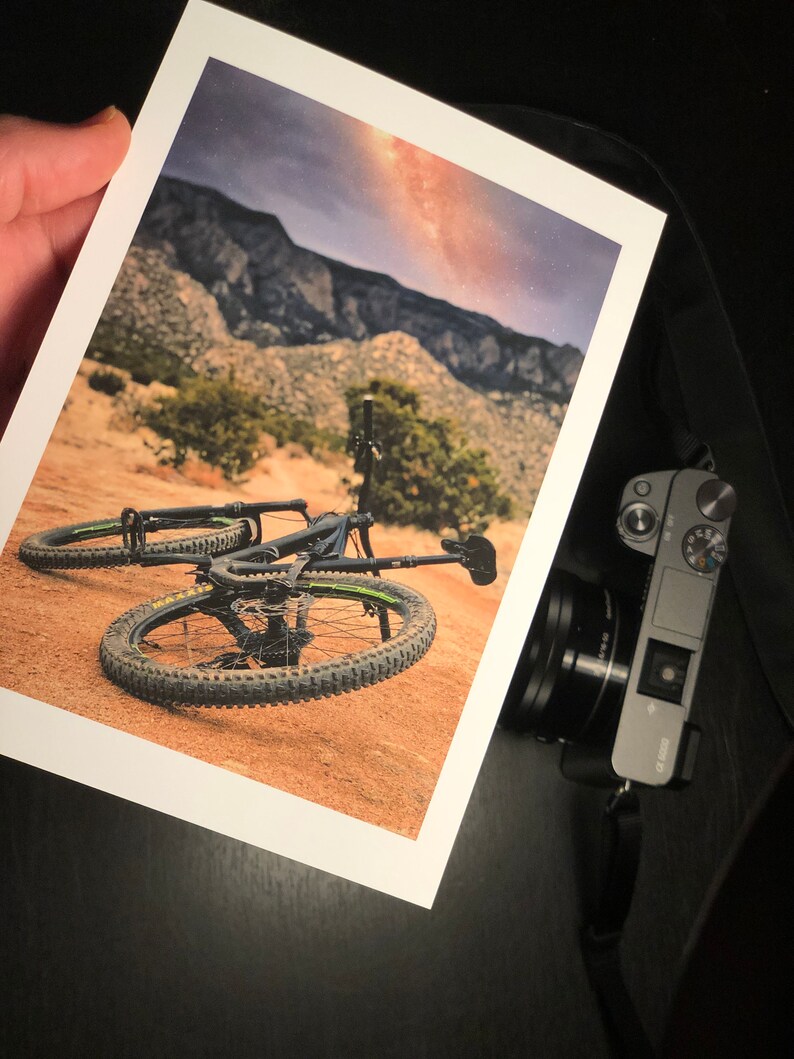 Pode incluir: Uma bicicleta de montanha preta com detalhes verdes est&aacute; deitada no ch&atilde;o em uma paisagem des&eacute;rtica. A bicicleta tem suspens&atilde;o dianteira e pneus com cravos. O fundo mostra uma cordilheira e um c&eacute;u nublado com um toque da Via L&aacute;ctea.