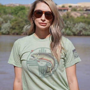 May include: A light green t-shirt with a graphic of a fish jumping out of the water. The fish is brown, orange, and yellow. The graphic is surrounded by a circle with a design that looks like a cross. The t-shirt has a small logo on the right sleeve that says "TRAV".