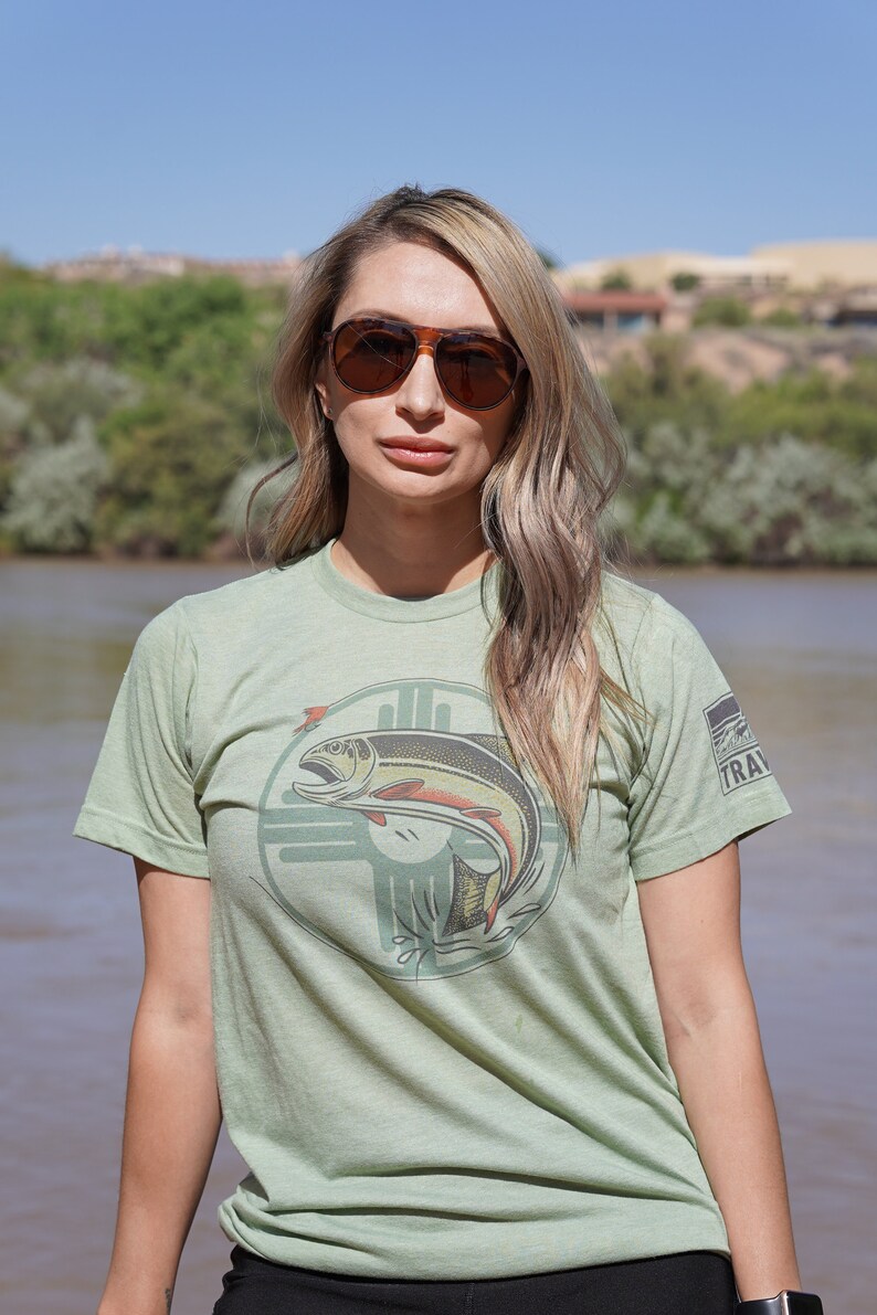 May include: A light green t-shirt with a graphic of a fish jumping out of the water. The fish is brown, orange, and yellow. The graphic is surrounded by a circle with a design that looks like a cross. The t-shirt has a small logo on the right sleeve that says "TRAV".