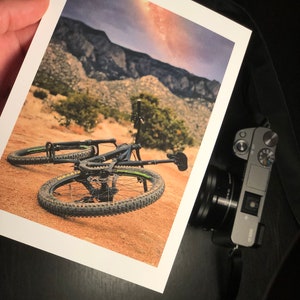 Pode incluir: Uma bicicleta de montanha preta com detalhes verdes est&aacute; deitada no ch&atilde;o em uma paisagem des&eacute;rtica. A bicicleta tem suspens&atilde;o dianteira e pneus com cravos. O fundo mostra uma cordilheira e um c&eacute;u nublado com um toque da Via L&aacute;ctea.