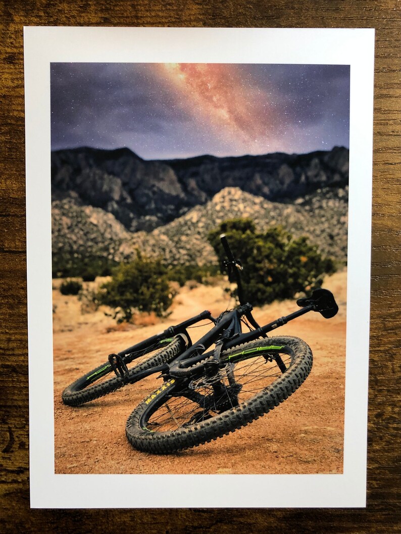 Pode incluir: Uma bicicleta de montanha preta com detalhes amarelos est&aacute; deitada de lado em um caminho de terra. A bicicleta est&aacute; na frente de uma cordilheira com um c&eacute;u estrelado acima.