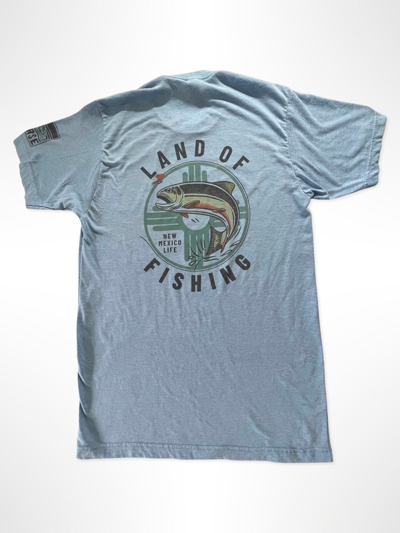 May include: A light blue t-shirt with a graphic of a fish jumping out of the water. The graphic is surrounded by a circle with the text "Land of Fishing" and "New Mexico Life".