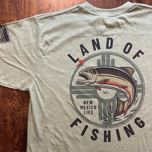 May include: A green t-shirt with a graphic of a fish jumping out of the water. The graphic is surrounded by a circle with the text "LAND OF FISHING" and "NEW MEXICO LIFE" inside.