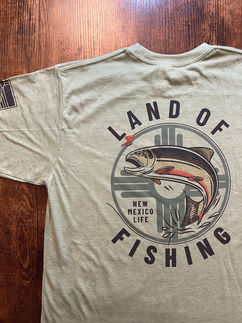 May include: A green t-shirt with a graphic of a fish jumping out of the water. The graphic is surrounded by a circle with the text "LAND OF FISHING" and "NEW MEXICO LIFE" inside.
