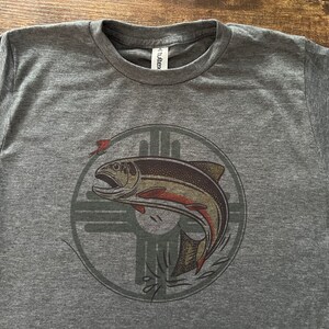 May include: A heather grey t-shirt with a graphic of a salmon jumping out of the water. The salmon is inside a circle with a stylized design of a cross. The design is in shades of brown, orange, and green.