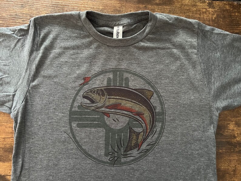 May include: A heather grey t-shirt with a graphic of a salmon jumping out of the water. The salmon is inside a circle with a stylized design of a cross. The design is in shades of brown, orange, and green.