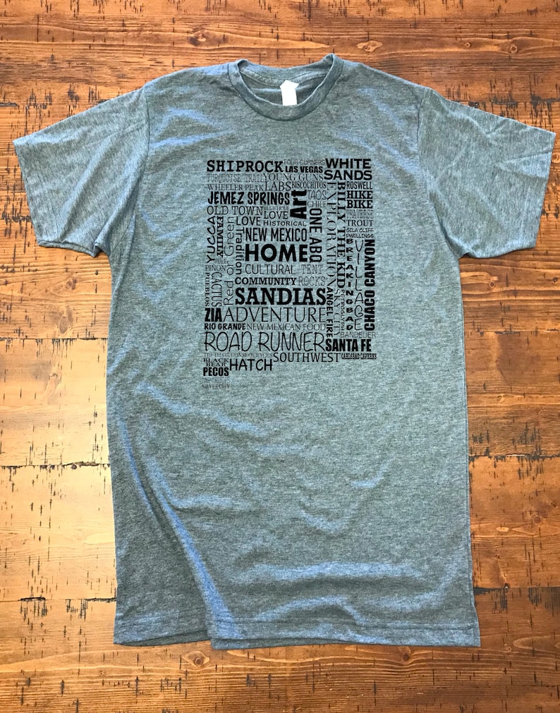 May include: A heather grey t-shirt with a black graphic print of a word cloud featuring places and things related to New Mexico. The words include "Shiprock", "Las Vegas", "White Sands", "Taos", "Jemez Springs", "Old Town", "Love", "New Mexico", "Home", "Cultural", "Community", "Sandals", "Adventure", "Rio Grande", "Road Runners", "Santa Fe", "Hatch", "Southwest", and more.