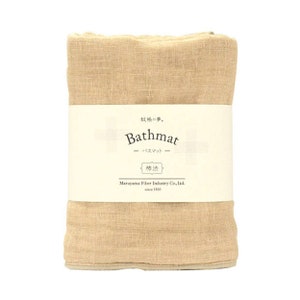 Puede incluir: Una alfombra de baño beige hecha de fibras naturales. La alfombra tiene una etiqueta que dice "Bathmat" en texto negro y "柿渋" en caracteres japoneses. La etiqueta también incluye el nombre de la empresa "Maruyama Fiber Industry Co., Ltd. since 1930".
