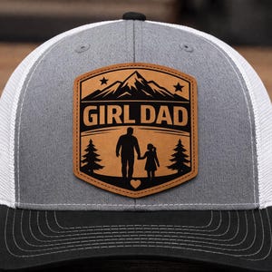 May include: A gray and white trucker hat with a black brim. The hat features a brown leather patch with the words "GIRL DAD" above a silhouette of a father and daughter walking, with a mountain and trees design.