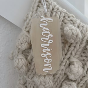 May include: A white acrylic tag with the word "Harrison" written in white script on a beige background. The tag is hanging from a white knitted stocking with gold thread accents.