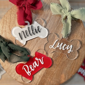 May include: Four clear acrylic dog bone ornaments with the names "Nellie", "Lucy", "Bear", and "Pelle" written in black. Each ornament is tied with a different colored ribbon.