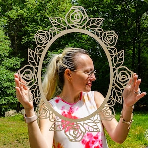 May include: A woman holds a wooden circular frame decorated with roses. The frame is made of light brown wood and has a delicate floral design. The woman is wearing a tie-dye shirt and has blonde hair.