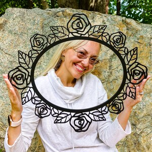 May include: A woman holds a black metal wreath with a rose design in front of her face. The wreath is circular and has a delicate floral pattern.