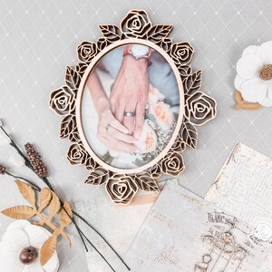 May include: Oval wooden picture frame with a laser-cut rose design. The frame holds a photo of two hands, one with a wedding ring. Decorative flowers and vintage paper accents surround the frame, creating a romantic theme.