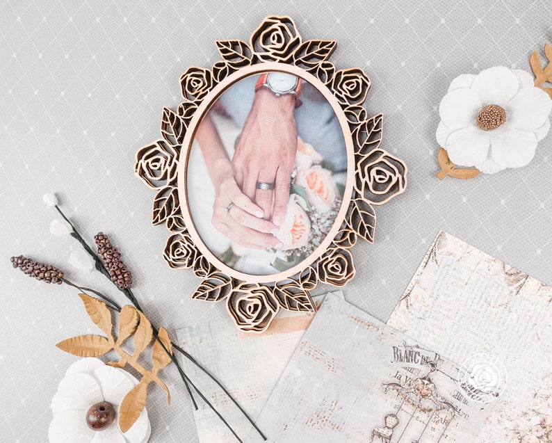 May include: Oval wooden picture frame with a laser-cut rose design. The frame holds a photo of two hands, one with a wedding ring. Decorative flowers and vintage paper accents surround the frame, creating a romantic theme.