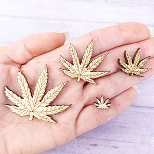 Puede incluir: Cuatro formas de hojas de cannabis de madera en varios tama&ntilde;os, mostradas en una mano. Las hojas son de color marr&oacute;n claro y tienen un patr&oacute;n de vetas detallado.