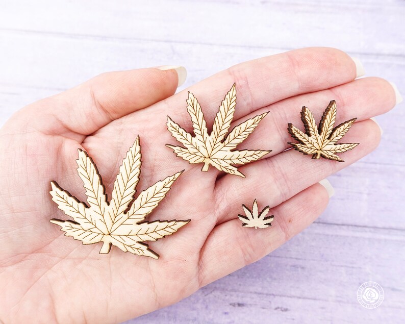 Puede incluir: Cuatro formas de hojas de cannabis de madera en varios tama&ntilde;os, mostradas en una mano. Las hojas son de color marr&oacute;n claro y tienen un patr&oacute;n de vetas detallado.