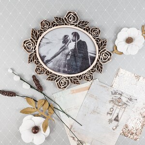 May include: A wooden oval picture frame with a rose design, holding a black and white photo of a couple kissing. Decorative paper, white flowers, and other floral accents are arranged around the frame on a gray patterned surface.