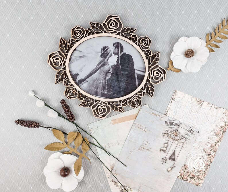 May include: A wooden oval picture frame with a rose design, holding a black and white photo of a couple kissing. Decorative paper, white flowers, and other floral accents are arranged around the frame on a gray patterned surface.