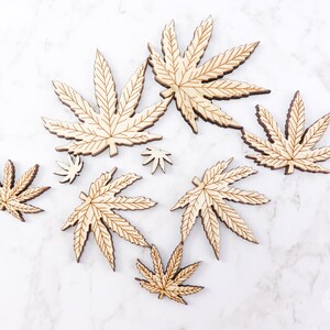 Puede incluir: Un conjunto de ocho formas de hojas de cannabis de madera cortadas con l&aacute;ser en varios tama&ntilde;os. Las hojas son de color marr&oacute;n claro y tienen un acabado de veta de madera natural.