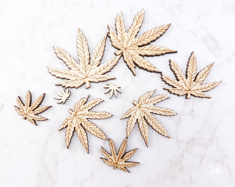 Puede incluir: Un conjunto de ocho formas de hojas de cannabis de madera cortadas con l&aacute;ser en varios tama&ntilde;os. Las hojas son de color marr&oacute;n claro y tienen un acabado de veta de madera natural.