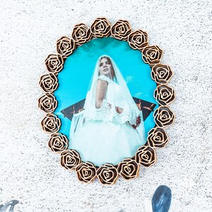 May include: A gold-coloured picture frame with a decorative rose design surrounds a photo of a woman in a white wedding dress. The photo is taken outdoors and the background is a blue sky.