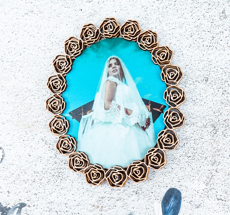 May include: A gold-coloured picture frame with a decorative rose design surrounds a photo of a woman in a white wedding dress. The photo is taken outdoors and the background is a blue sky.