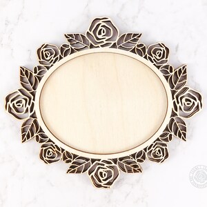 May include: A wooden oval frame with a floral rose design. The frame is made of light-colored wood with a laser-cut rose and leaf border. The center is a smooth, blank oval. The frame is approximately 10 inches wide.