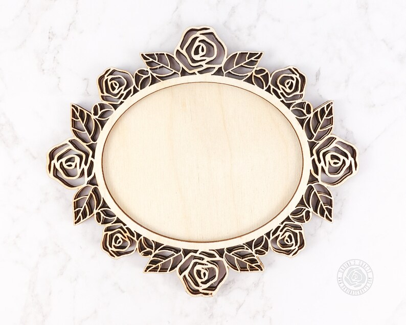 May include: A wooden oval frame with a floral rose design. The frame is made of light-colored wood with a laser-cut rose and leaf border. The center is a smooth, blank oval. The frame is approximately 10 inches wide.