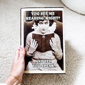 May include: A vintage-style book cover with a woman reading a book. The text on the cover reads "YOU SEE ME READING, RIGHT? AND YET, YOU SPEAK."