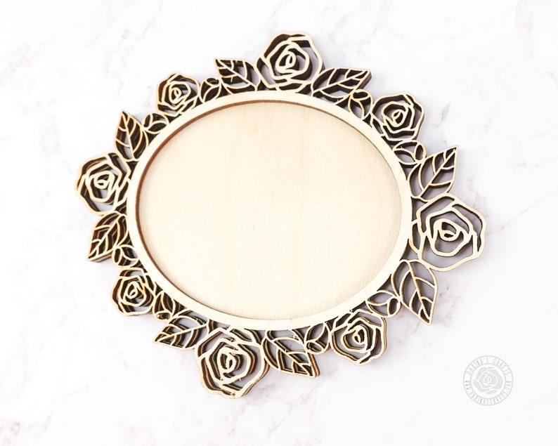 May include: A laser-cut wooden decorative piece featuring a central oval area surrounded by a floral design. The design includes roses and leaves, with a natural wood finish. The piece is likely intended for crafting or home decor.