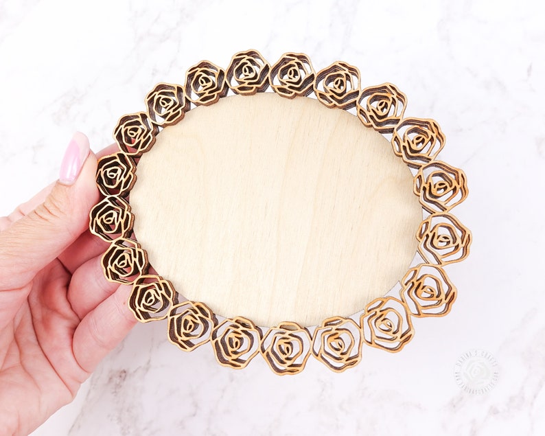 May include: A round wooden plaque with a decorative border of laser-cut roses. The plaque is light brown and the roses are a darker brown.